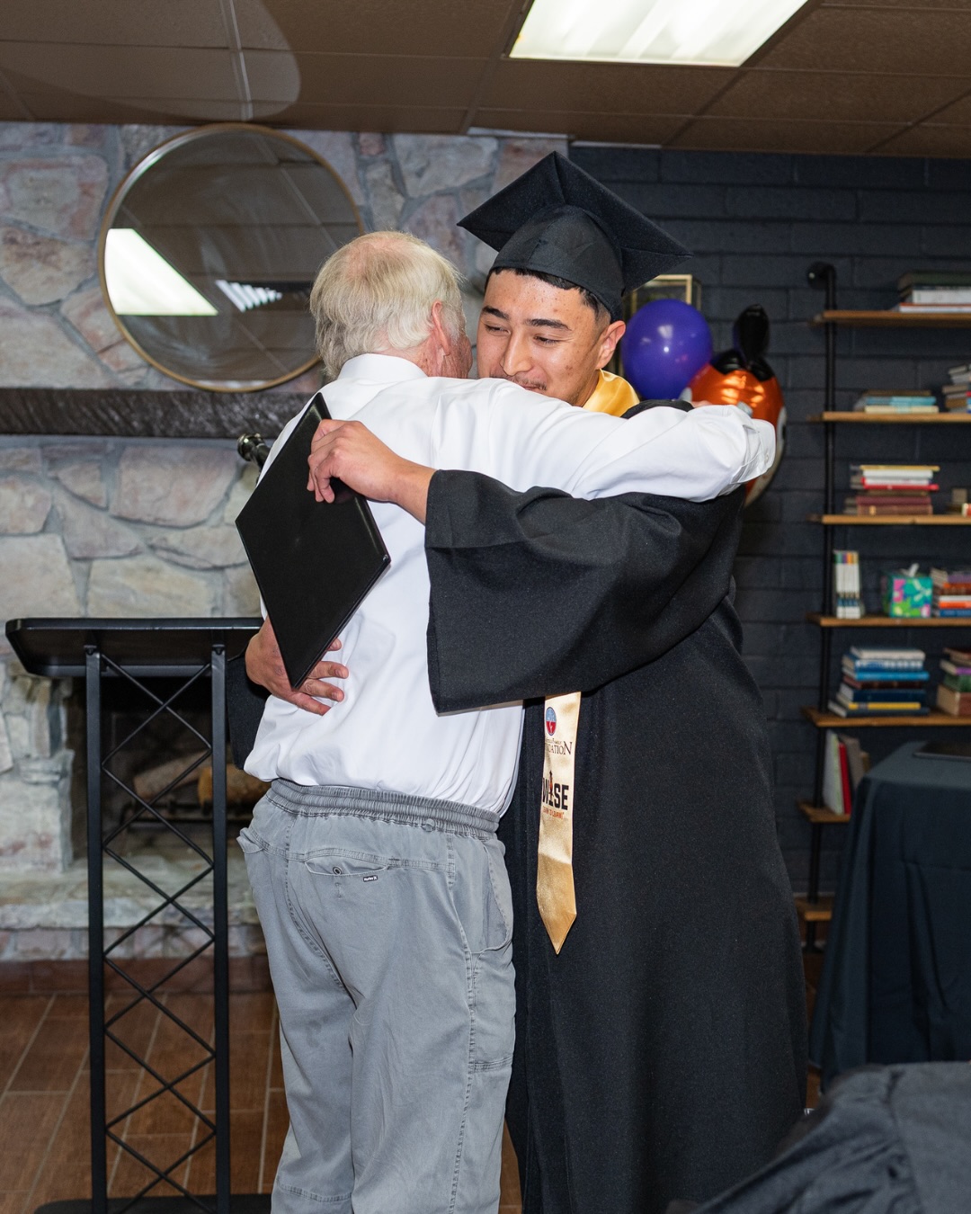 A graduate in a cap and gown embraces an attendee at the celebration.
